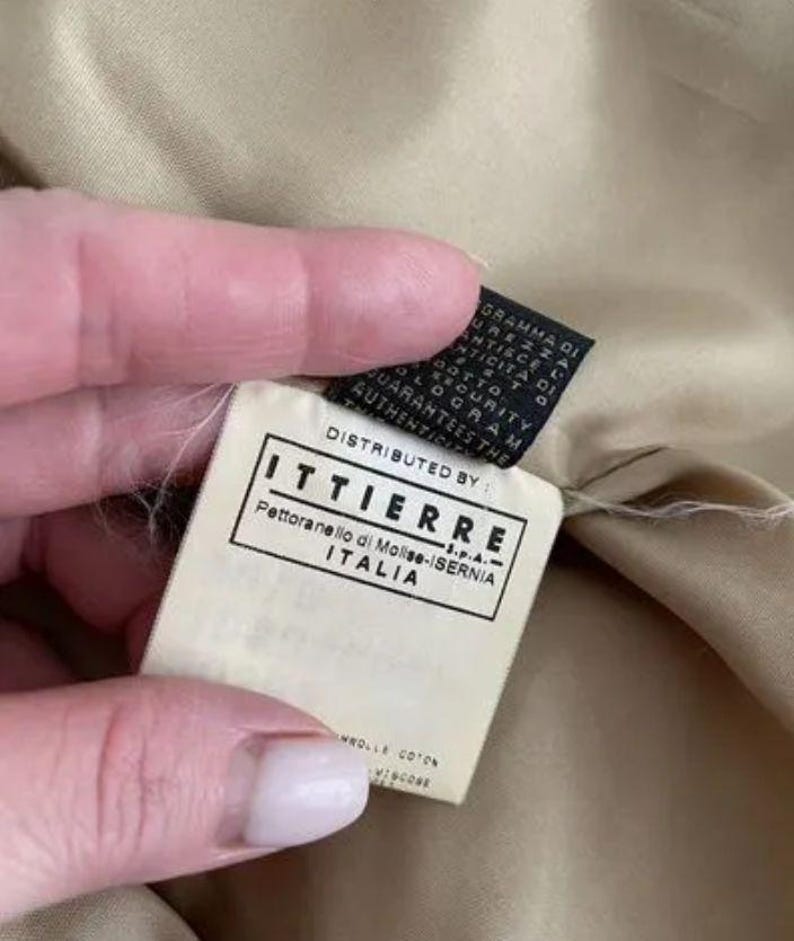 May include: A beige fabric garment interior featuring a white rectangular clothing label with black text that reads DISTRIBUTED BY ITTIERRE S.P.A. Pettoranello di Molise-ISERNIA ITALIA, positioned above a smaller black security tag.