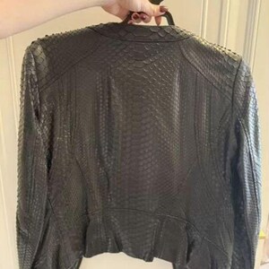 May include: A black jacket featuring a textured snakeskin-like pattern across the entire surface. The garment has long sleeves, a rounded neckline, and a structured fit with visible seam detailing along the back and shoulders.