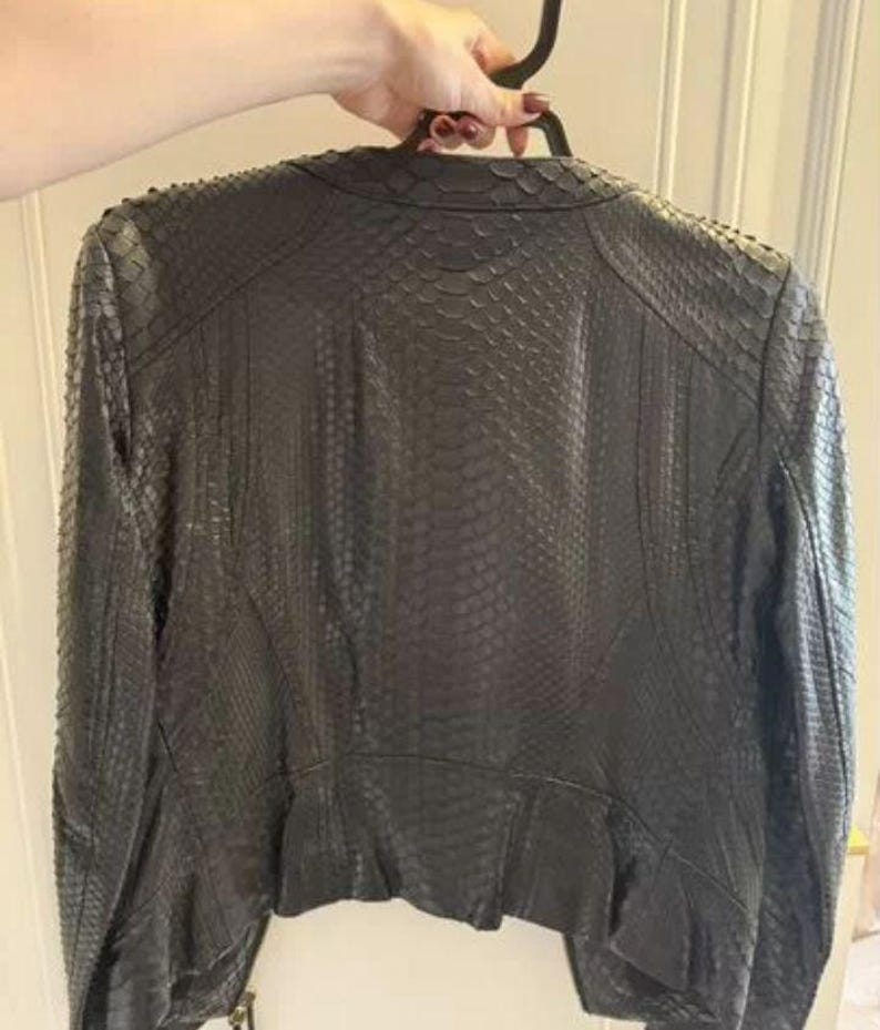 May include: A black jacket featuring a textured snakeskin-like pattern across the entire surface. The garment has long sleeves, a rounded neckline, and a structured fit with visible seam detailing along the back and shoulders.