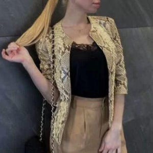 May include: A beige and brown snakeskin print jacket with three-quarter length sleeves and a collarless neckline. The jacket is worn over a black camisole with lace trim and paired with tan trousers. A black handbag with a gold chain strap is visible.