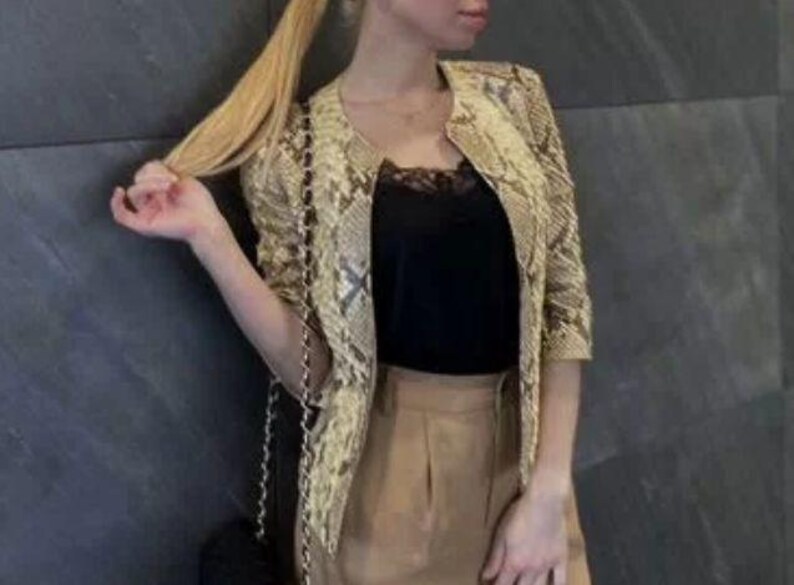 May include: A beige and brown snakeskin print jacket with three-quarter length sleeves and a collarless neckline. The jacket is worn over a black camisole with lace trim and paired with tan trousers. A black handbag with a gold chain strap is visible.