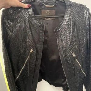 May include: A black jacket featuring a textured snakeskin pattern throughout the exterior. The garment has a round neckline, long sleeves, and two diagonal zippered pockets on the front. It includes a dark interior lining and a brand label.