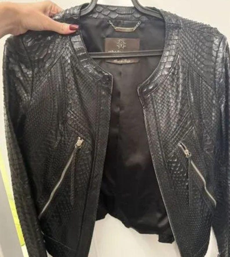 May include: A black jacket featuring a textured snakeskin pattern throughout the exterior. The garment has a round neckline, long sleeves, and two diagonal zippered pockets on the front. It includes a dark interior lining and a brand label.