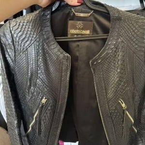 May include: A black jacket with a textured snakeskin pattern, featuring a round neckline, long sleeves, and two front pockets with gold-tone metal zippers. The interior is lined with black fabric and includes a Roberto Cavalli brand label.