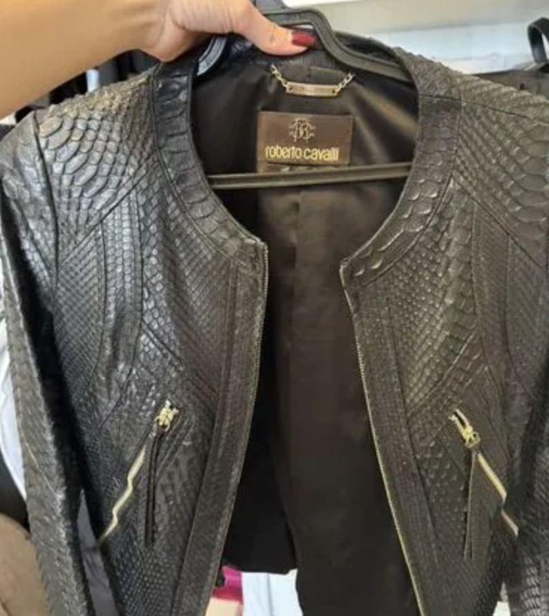 May include: A black jacket with a textured snakeskin pattern, featuring a round neckline, long sleeves, and two front pockets with gold-tone metal zippers. The interior is lined with black fabric and includes a Roberto Cavalli brand label.
