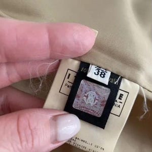 May include: A beige garment interior featuring a white fabric tag with the number 38 printed in black. A black rectangular label is attached, displaying a holographic square emblem with a logo and the text BAUMWOLLE COTON VISKOSE VISCOSE.