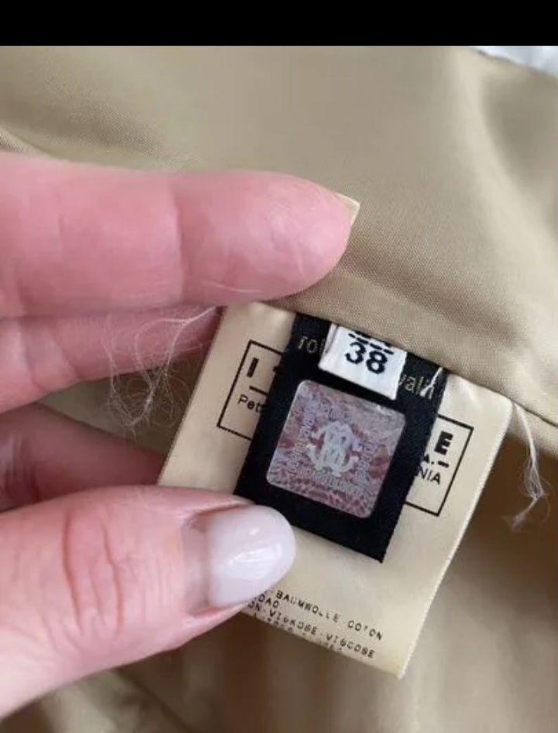 May include: A beige garment interior featuring a white fabric tag with the number 38 printed in black. A black rectangular label is attached, displaying a holographic square emblem with a logo and the text BAUMWOLLE COTON VISKOSE VISCOSE.
