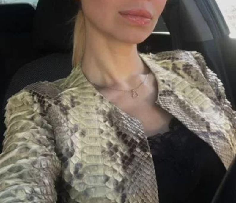 May include: A jacket featuring a textured snakeskin pattern in shades of beige, cream, and dark brown. The garment has a structured collarless neckline and is worn over a black top with lace trim. A thin gold chain necklace with a small pendant is visible.