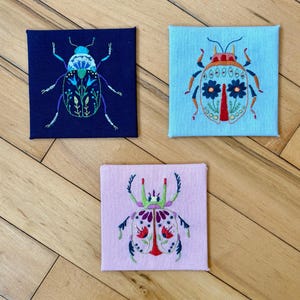May include: Three square embroidered canvases featuring colorful beetle designs. One is on a navy blue background, another on light blue, and the third on pink. The beetles are decorated with floral and geometric patterns, showcasing a handcrafted aesthetic.