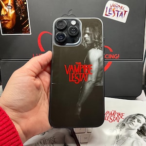 May include: Black phone case featuring the text "THE VAMPIRE LESTAT" in red. The case has a black and white image of a man. A sticker with the text "THE VAMPIRE LESTAT" is also visible.