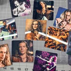 May include: A collection of square coasters featuring images of actors and text from the movie "Vampire Lestat". The coasters display various portraits and scenes, with a dominant color palette of purple, black, and orange.