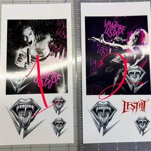 May include: Two rectangular sticker sheets featuring The Vampire Lestat imagery. Sheet one shows a person singing with the text The Vampire Lestat and three silver vampire mouth icons. Sheet two shows a person performing with the text The Vampire Lestat, the word Lestat in red, and two silver vampire mouth icons.