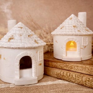 May include: Two white ceramic house-shaped incense burners with textured roofs and gold-colored flecks. One house features a chimney emitting smoke, while the other contains a glowing yellow light inside its arched doorway and a small window.
