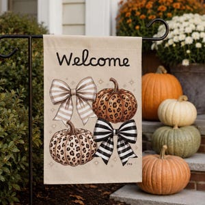 May include: A rectangular garden flag with a beige background featuring the word Welcome in black script. The design includes two pumpkins with brown leopard print patterns, one beige and white striped bow, and one black and white striped bow.