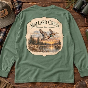 May include: A sage green long-sleeve shirt featuring a large graphic on the back. The design shows two mallard ducks flying over a river at sunset, surrounded by pine trees, with the text "MALLARD CREEK" and "Newland Row Outdoors" printed above.