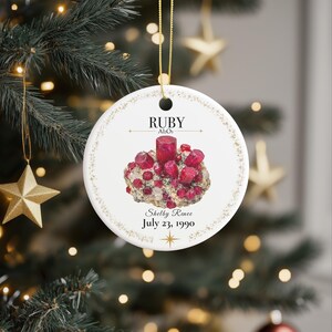May include: A round white ceramic ornament featuring a central illustration of a red ruby crystal cluster. The text reads RUBY, Al2O3, Shelby Renee, and July 23, 1990. The design includes a gold star icon and a delicate gold border, suspended by a gold cord.