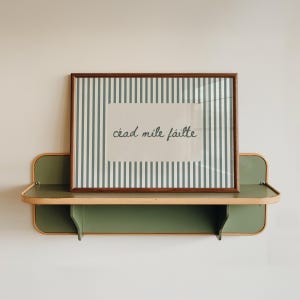 May include: A framed print with vertical green and white stripes and the text "céad míle fáilte" in a handwritten style. The print is displayed on a green shelf with a curved design, mounted on a white wall.