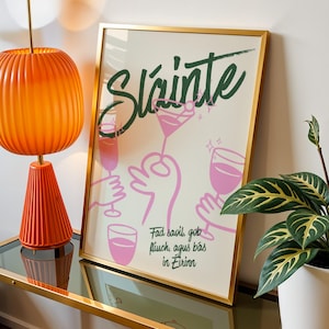 Könnte beinhalten: Ein gerahmter Kunstdruck mit goldenem Rahmen zeigt das Wort "Sláinte" in grüner Schrift, mit rosa Illustrationen von Personen, die mit Getränken anstoßen. Der Druck enthält auch Text in kleinerer Schrift. Eine orangefarbene Lampe und eine Topfpflanze sind ebenfalls zu sehen.