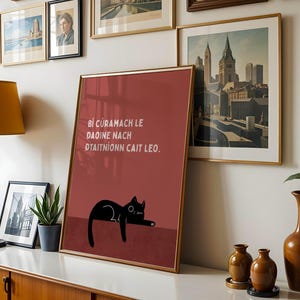 May include: A framed art print featuring a black cat illustration on a burgundy background. The cat is lying down with its eyes closed. The print includes text in white. The artwork is displayed on a white surface with other framed art and decor.