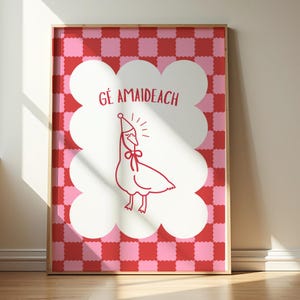 May include: A framed art print featuring a red and pink checkered background. A white cloud-shaped design contains a red illustration of a goose wearing a party hat and a bow. The text "GÉ AMAIDEACH" is above the goose.