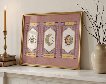 May include: A framed print featuring three tarot card designs. Each card has a central image: an eye, a crescent moon, and a sun with a face. The cards are set against a pink background with gold and purple accents. The frame is gold.