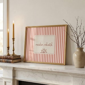May include: A rectangular gold-framed print featuring vertical pink and white stripes with a central cream-colored rectangle containing the text "maidin mhaith" in brown script above a small brown sun icon, displayed in a home interior setting.