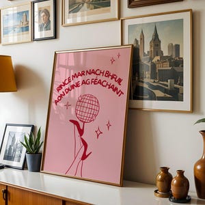 May include: A rectangular framed wall art print featuring a pink background with dark red line art of a disco ball and high-heeled legs. The text reads: RINCE MAR NACH BHFUIL AON DUINE AG FÉACHAINT. Four small four-pointed star icons surround the central graphic.