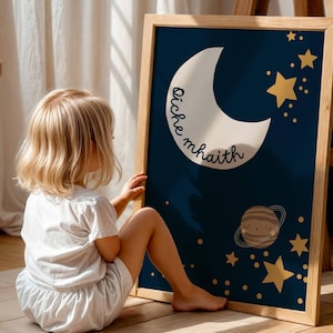 May include: A framed art print with a navy blue backdrop, a white crescent moon, and gold stars. The print includes the text "oiche mhaith" and a planet illustration. The frame is made of light-coloured wood.