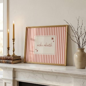 May include: A rectangular gold-framed print featuring a vertical pink and white striped pattern. The center contains a cream-colored rectangular label with the text "oíche mhaith" in a dark script font, accented by small star-shaped icons.