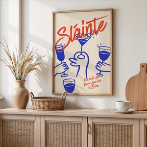 Può includere: Una stampa artistica incorniciata con sfondo crema presenta la parola "Sláinte" in corsivo rosso sopra illustrazioni blu di mani che tengono bicchieri da cocktail. La stampa include testo in un carattere più piccolo. L'opera d'arte è esposta su un mobile in legno.
