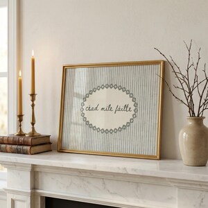 May include: A framed artwork with a gold-colored frame. The artwork features a striped background with a central oval design and the text "ciad mile fáilte". Two lit candles in brass holders and a vase with branches are also visible.