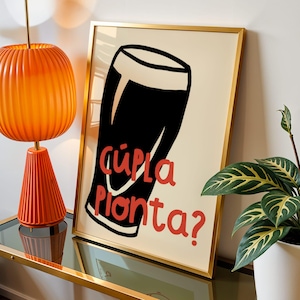 May include: A framed wall art print featuring a stylised black and white illustration of a pint glass with a creamy head. The text "Cúpla Pionta?" is printed in red lettering across the centre of the glass against a cream-coloured background.