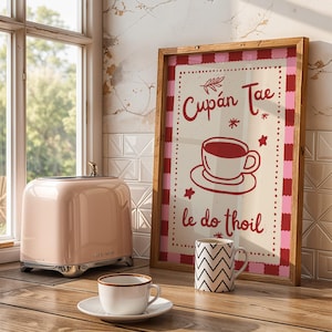 Op de afbeelding: Een ingelijste wandprint met een rood en roze geblokte rand, een centrale illustratie van een theekopje op een schoteltje en de rode tekst Cupán Tae le do thoil. De print staat op een houten oppervlak naast een roze broodrooster en twee mokken.