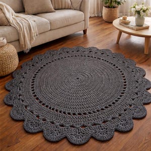 Könnte beinhalten: Ein dunkelgrauer, runder Häkelteppich mit einem gewellten Rand. Der Teppich liegt auf einem Holzboden in einem Wohnzimmer. Der Durchmesser des Teppichs beträgt ca. 152 cm.