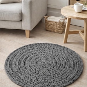 Könnte beinhalten: Ein runder, grauer gehäkelter Teppich mit Spiralmuster. Der Teppich liegt auf einem hellen Holzboden, mit einem Sofa, einem Beistelltisch und einem Korb im Hintergrund. Der Teppich hat einen Durchmesser von etwa 100 cm.