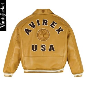 May include: Mustard yellow leather jacket with the word "AVIREX" arched across the back, above "USA". A circular emblem is centered between the words. The jacket has a collared neckline and ribbed cuffs with white stripes.
