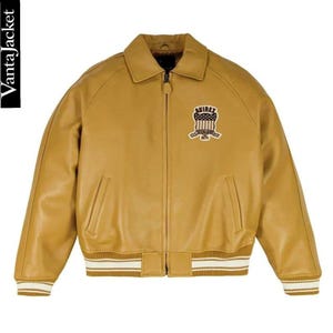 May include: Mustard-colored leather jacket with a zip-up front, collar, and ribbed cuffs and hem. Features a patch on the chest with the text "AVIREX" and an emblem. The jacket has two front pockets and the text "VantaJacket" on the side.