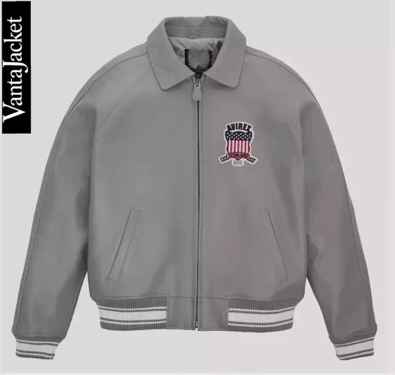 May include: A gray bomber jacket with a collared neckline and a zipper closure. The jacket features a patch on the chest with the text "AVIREX" and a design in red, white, and blue. The cuffs and hem have white and gray striped detailing. The text "VantaJacket" is on the left side.