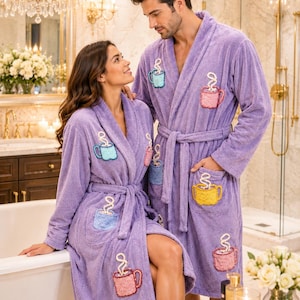 May include: Two people wearing matching lavender bathrobes with embroidered coffee cup designs. The robes have a shawl collar, pockets, and a waist tie. The bathroom setting includes a bathtub, chandelier, and gold fixtures.