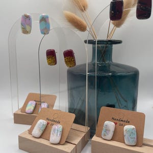 May include: A display of handmade stud earrings in various designs. Some earrings feature marbled patterns in pastel colors, while others have a glittery gold or burgundy finish. The earrings are presented on wooden stands and clear acrylic arches.