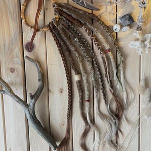 May include: A collection of handmade dreadlocks in various shades of brown, beige, and gray, with decorative wraps and beads. The dreadlocks are arranged against a wooden background, showcasing a bohemian or natural aesthetic.