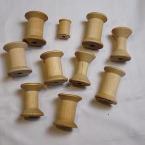 May include: A collection of wooden spools in various sizes, arranged on a white surface. The spools are cylindrical with flared ends, showcasing the natural wood grain. Ideal for crafts and sewing projects.