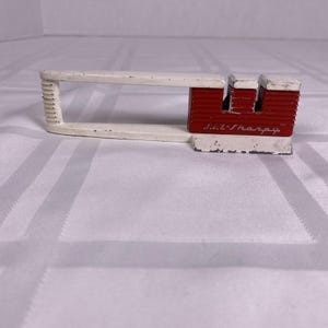 May include: A vintage knife sharpener with a white handle and a red sharpening block. The handle has a rectangular shape with a ribbed grip. The red block has two slots for sharpening and the text "Lil' Sharp" is printed on the side.