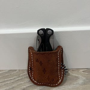 May include: A brown leather multi-tool case with a white stitched border. The case holds a black and silver multi-tool. The case has a diamond pattern and a black and white cord attached.