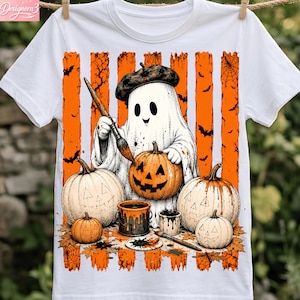 Op de afbeelding: Een wit t-shirt met een afbeelding van een geest met een baret die een pompoen beschildert. Het ontwerp bevat oranje verticale strepen, vleermuizen, spinnenwebben, meerdere pompoenen, verfblikken en penselen op een witte achtergrond.