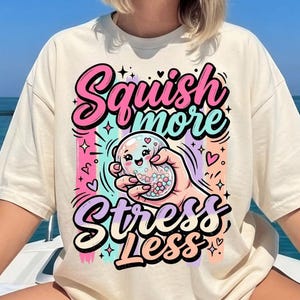 May include: Cream-coloured t-shirt featuring the text "Squish More Stress Less" in a colourful, playful design. The graphic shows a hand holding a cute, smiling squishy toy with heart and star accents. The shirt is worn by a person.