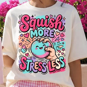 Può includere: Una t-shirt a maniche corte color crema con una stampa grafica colorata. Il design mostra una mano che stringe una pallina antistress blu sorridente, circondata da cuori e stelle rosa, con il testo "Squish More Stress Less" in grassetto.