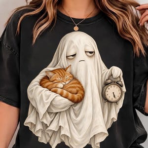 Könnte beinhalten: Ein schwarzes T-Shirt mit einer Grafik eines weißen Geistes mit müdem Gesichtsausdruck, der eine schlafende orangefarbene getigerte Katze im Arm hält und einen kleinen, runden, dunkel gefärbten analogen Wecker in der anderen Hand trägt.