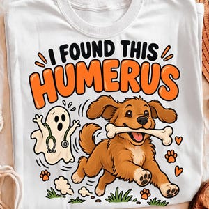 May include: A white t-shirt featuring a graphic of a brown dog running with a bone in its mouth next to a small ghost wearing a stethoscope. The text reads I FOUND THIS HUMERUS in black and orange lettering, surrounded by small paw prints and hearts.
