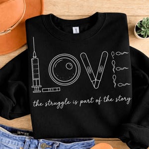 Op de afbeelding: Zwart sweatshirt met een wit grafisch ontwerp. Het woord "LOVE" is gevormd met een spuit, een ei, zwangerschapstests en sperma-illustraties. De tekst "the struggle is part of the story" staat eronder.